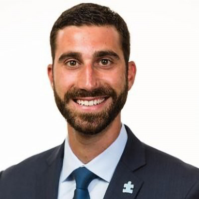 The image shows a man in a suit and tie. He has short dark hair, a beard, and is smiling at the camera. He appears to be in a professional setting. There is a small pin on his lapel. The background is plain white.